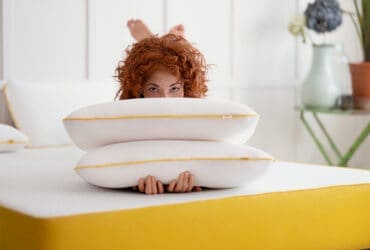 Pillow stack on bed with woman hiding behind it, cozy home bedroom decor.