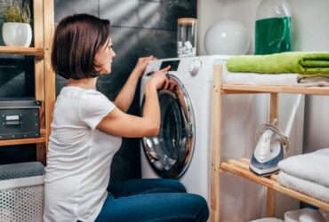 A woman operating a washing machine in a laundry room with shelves and cleaning supplies.